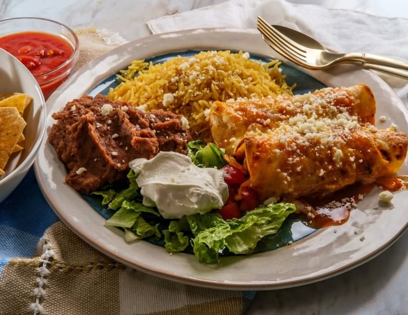 Two enchiladas with rice, refried beans, lettuce, sour cream, and salsa on a plate.