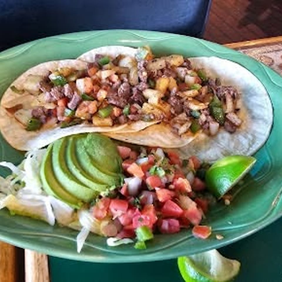 Two street tacos with meat, potatoes, and peppers, served with avocado, pico de gallo, and lime.