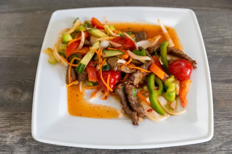 Spicy beef salad with sliced vegetables like bell peppers, onions, and carrots on a white plate.