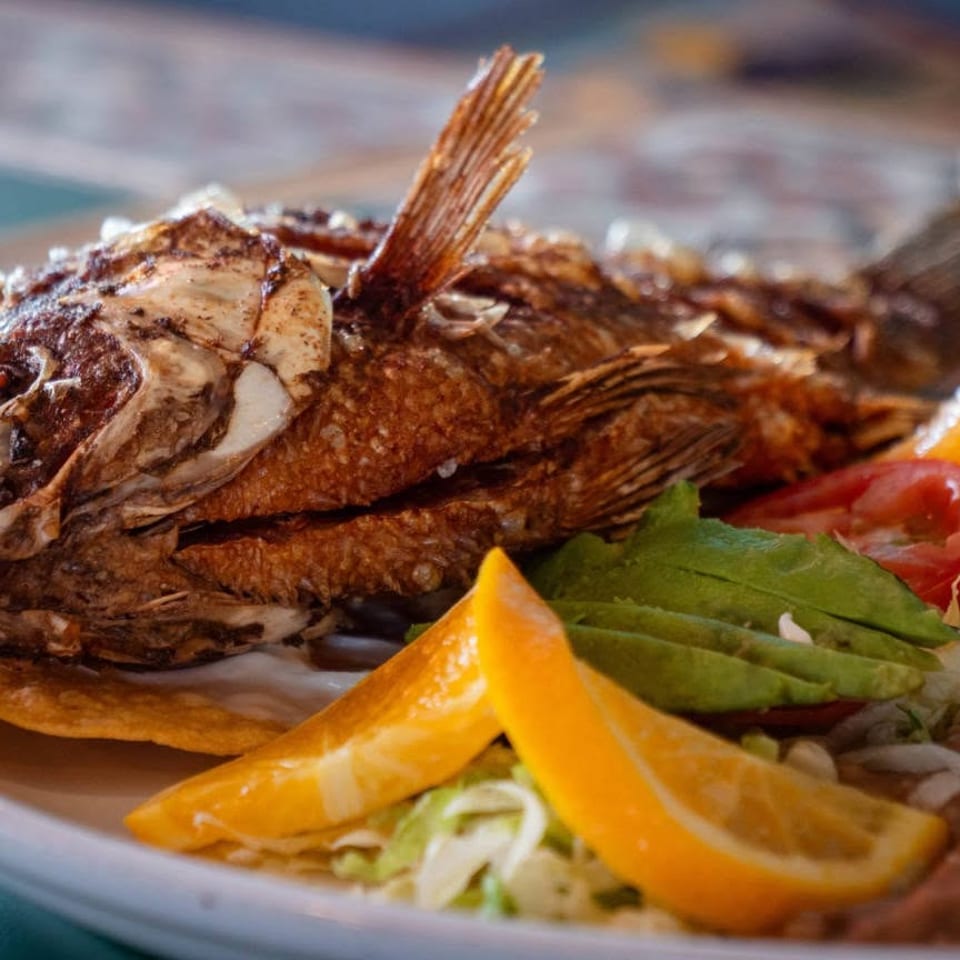 Whole fried fish served with avocado, tomato, lettuce, refried beans, and orange slices.