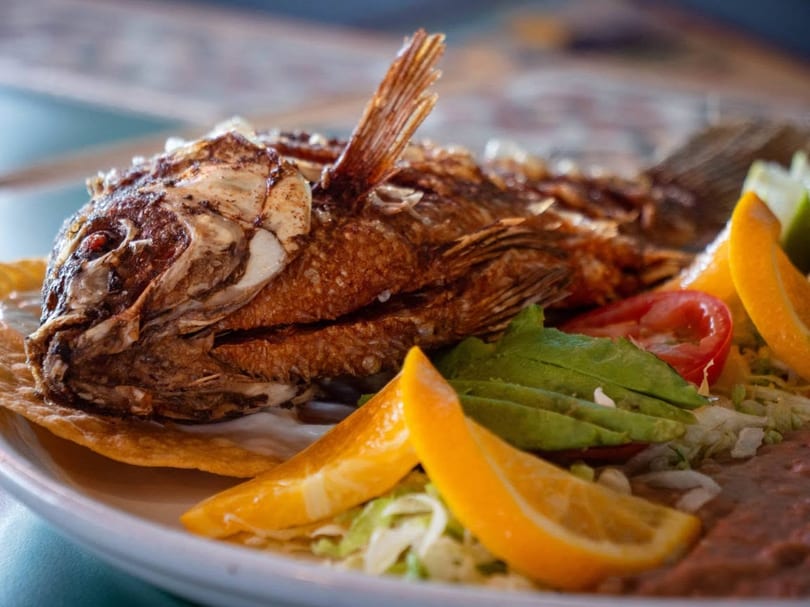 Whole fried fish served with avocado, tomato, lettuce, refried beans, and orange slices.