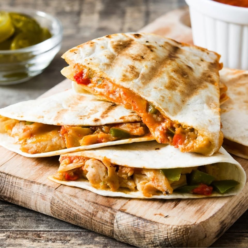Chicken and vegetable quesadillas on a wooden board, with salsa and pickled jalapeños.