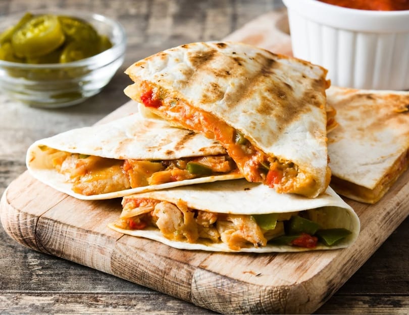 Chicken and vegetable quesadillas on a wooden board, with salsa and pickled jalapeños.