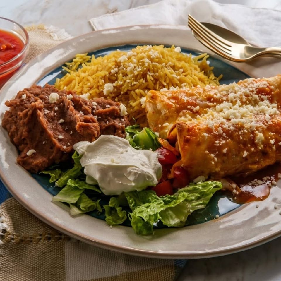 Two enchiladas topped with cheese, served with yellow rice, refried beans, lettuce, and sour cream.
